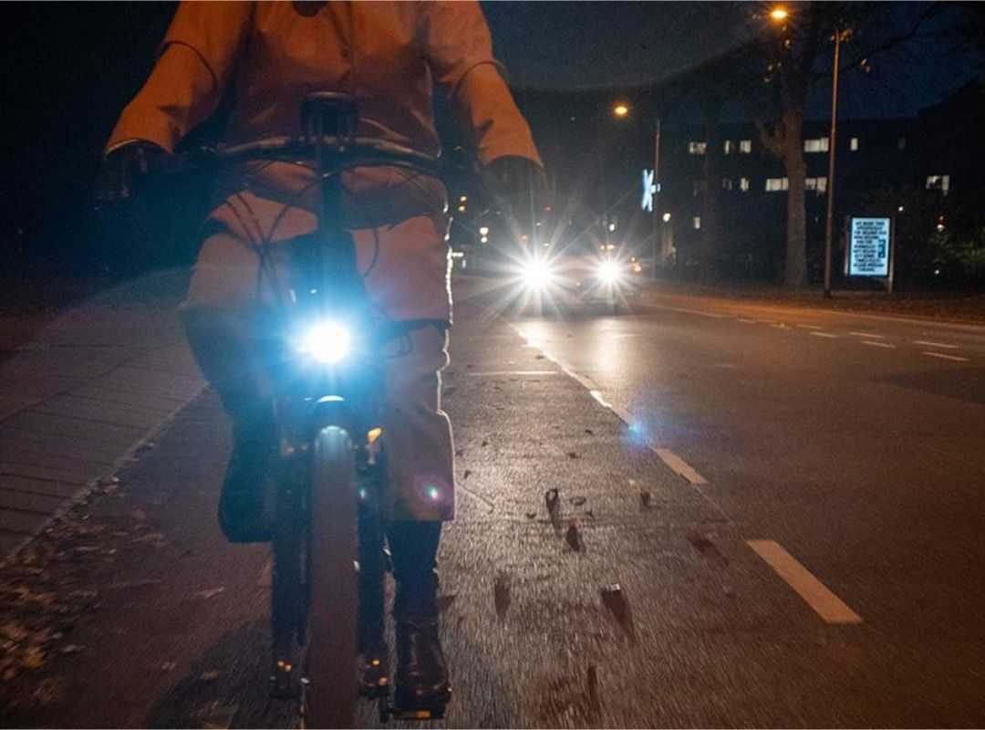 Person riding an e-bike at night with a bright front light illuminated, traveling along a city street as approaching vehicle headlights shine in the background.